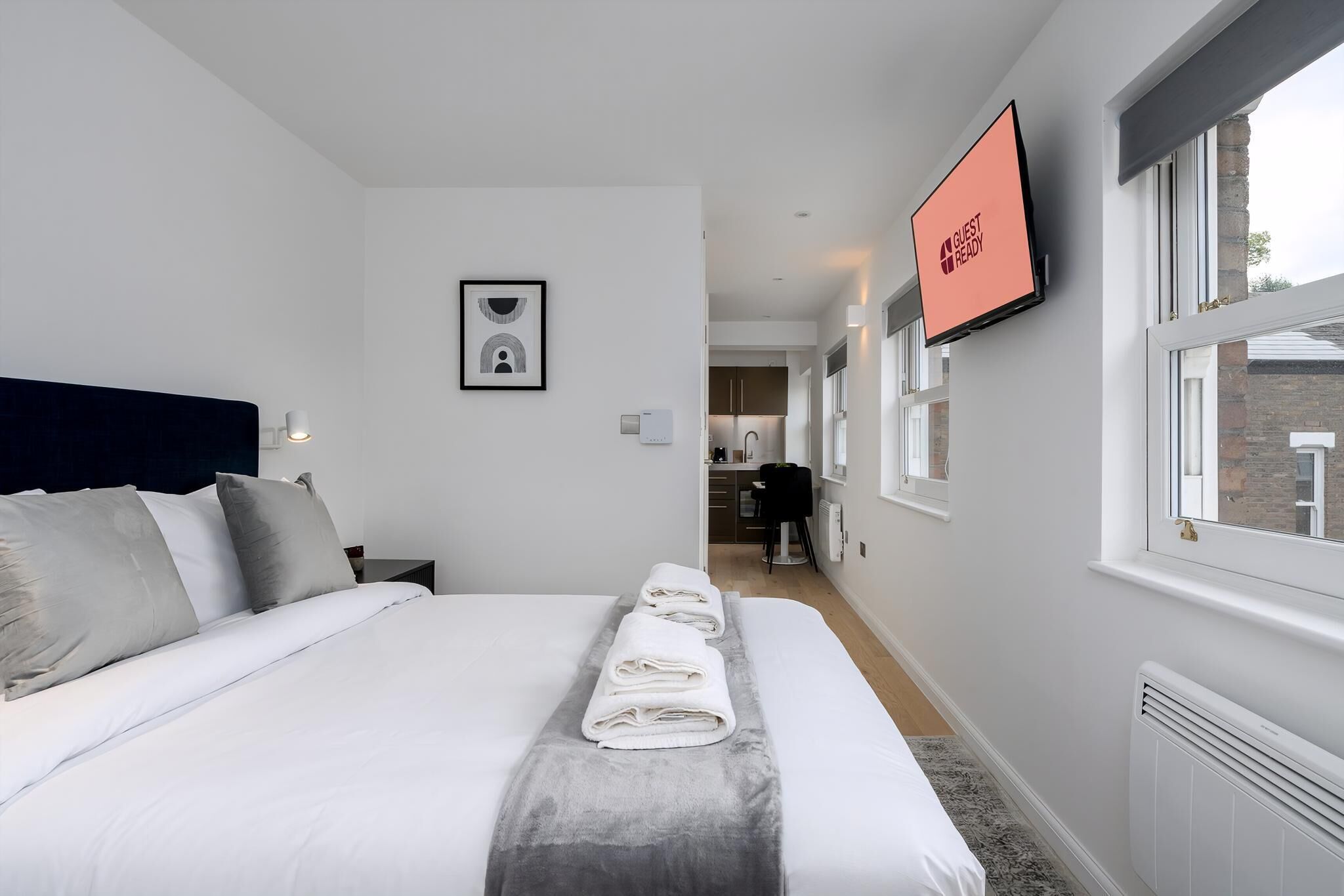 A hotel room with a neatly made bed, a flat-screen TV on the wall, and a view of the kitchen area.