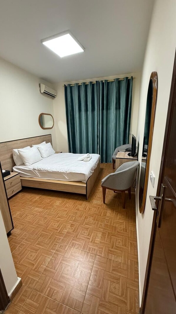 A hotel room with a bed, a desk, a chair, a mirror, an air conditioning unit, and a window with curtains. - Weekend Hotel (Beirut)