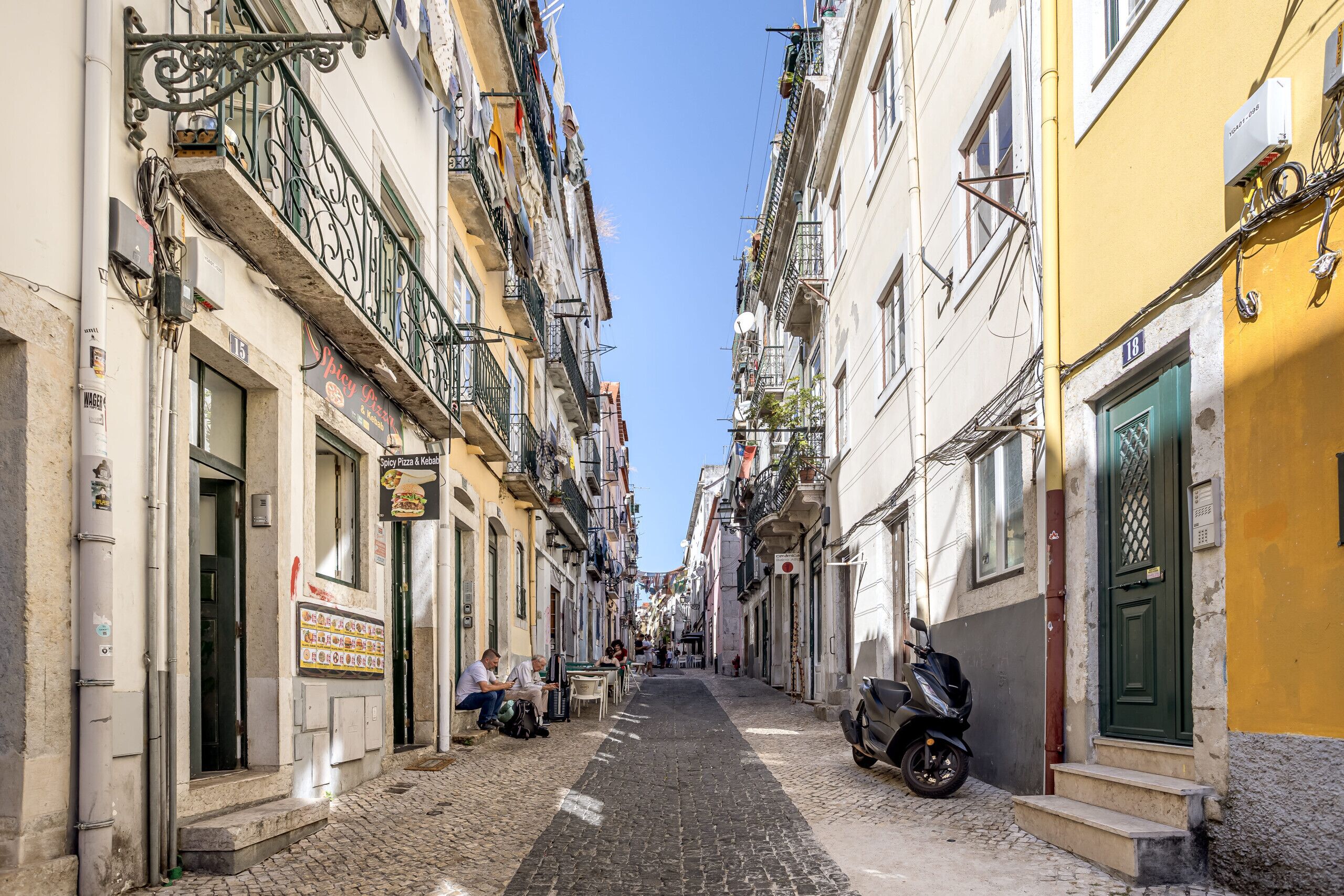 A narrow cobblestone street flanked by buildings with balconies and outdoor seating areas.