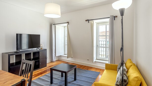 A modern living room with a flat-screen TV, wooden dining table, yellow sofa, and large windows with curtains. - Bairro Alto Typical by Homing (Lisbon)