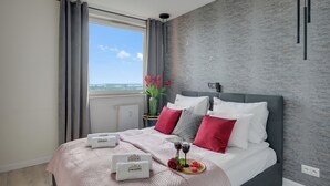 A modern hotel room with a large bed, grey and white bedding, a view of the city, and a vase with flowers.