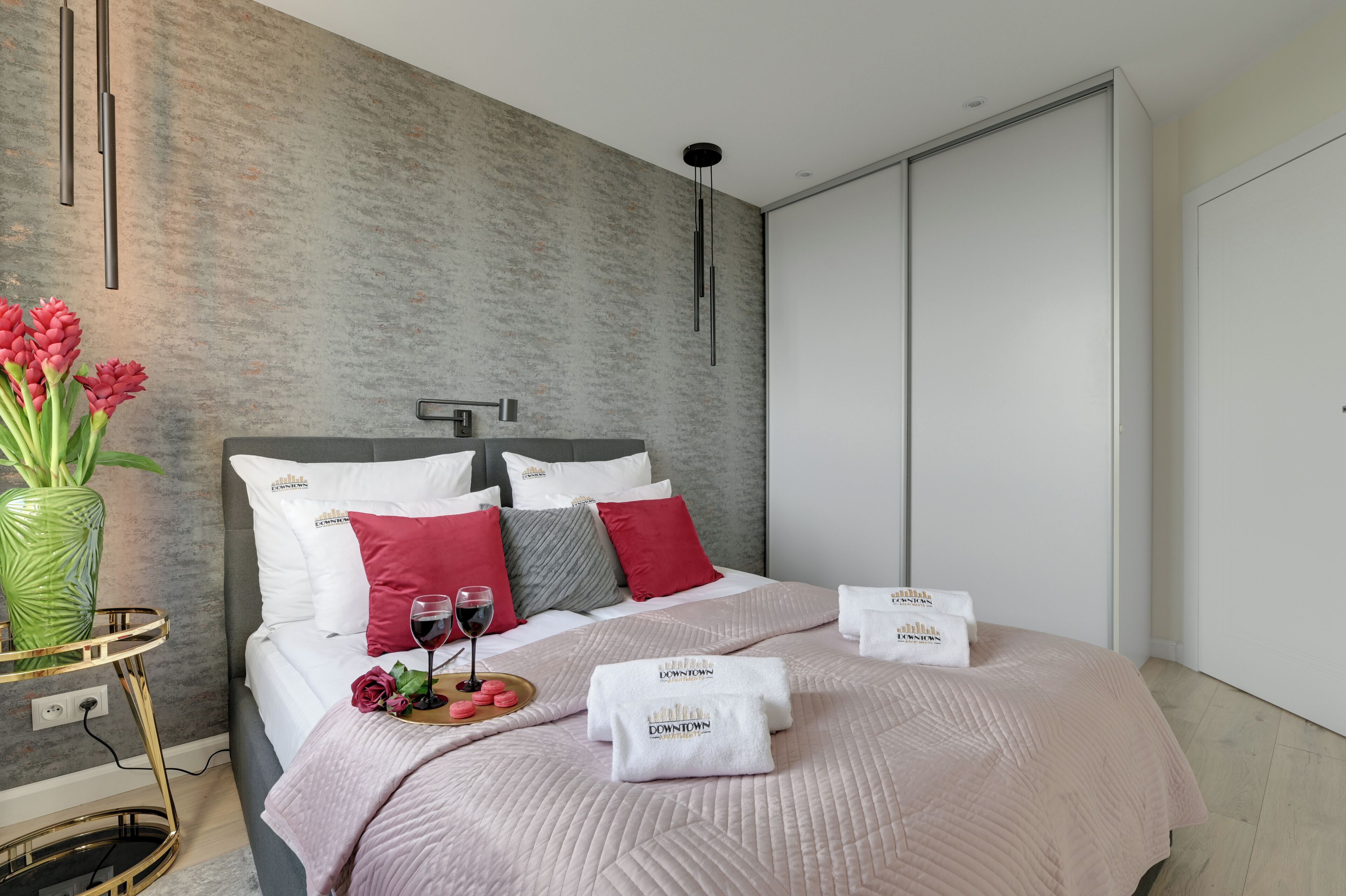 Un dormitorio moderno con una cama, almohadas, un florero con flores, una bandeja con aperitivos y una copa de vino.