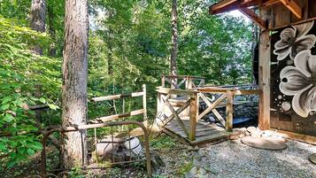 Une terrasse en bois rustique, dotée d’une rambarde en métal, entourée d’arbres et bordée d’un chemin de gravier.