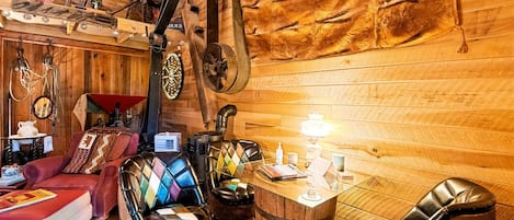 Une pièce rustique avec des murs en bois, un plafond en bois, un canapé rouge, deux fauteuils en cuir, un tonneau en bois et une machine à coudre ancienne.