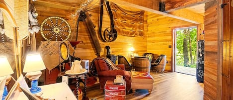 A rustic cabin interior with wooden walls, a wooden ceiling, a sofa, a table with a lamp, and a large window.