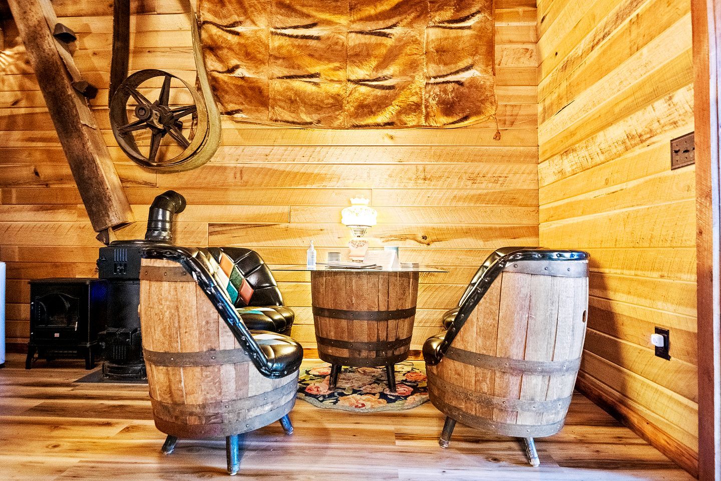A rustic interior with wooden walls, a large fur rug, a wooden barrel-shaped table, and a vintage stove.