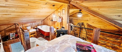 Un dormitorio en ático con techo inclinado, una bañera de patas de león roja, una cama con ropa de cama estampada y una pequeña mesa con una silla.