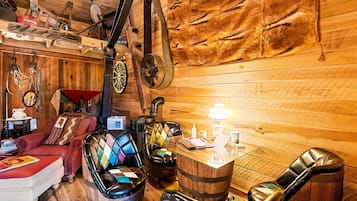 A rustic room with wooden walls, a wooden ceiling, a red sofa, two leather armchairs, a wooden barrel, and a vintage sewing machine.