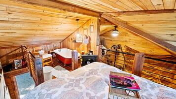 A wooden cabin bedroom with a sloped ceiling, a red clawfoot bathtub, a bed with patterned bedding, and a small table with a lamp.