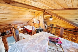 A wooden cabin bedroom with a sloped ceiling, a red clawfoot bathtub, a bed with patterned bedding, and a small table with a lamp.