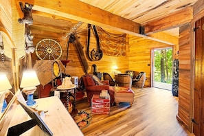 A rustic cabin interior with wooden walls, a wooden ceiling, a sofa, a table with a lamp, and a large window.