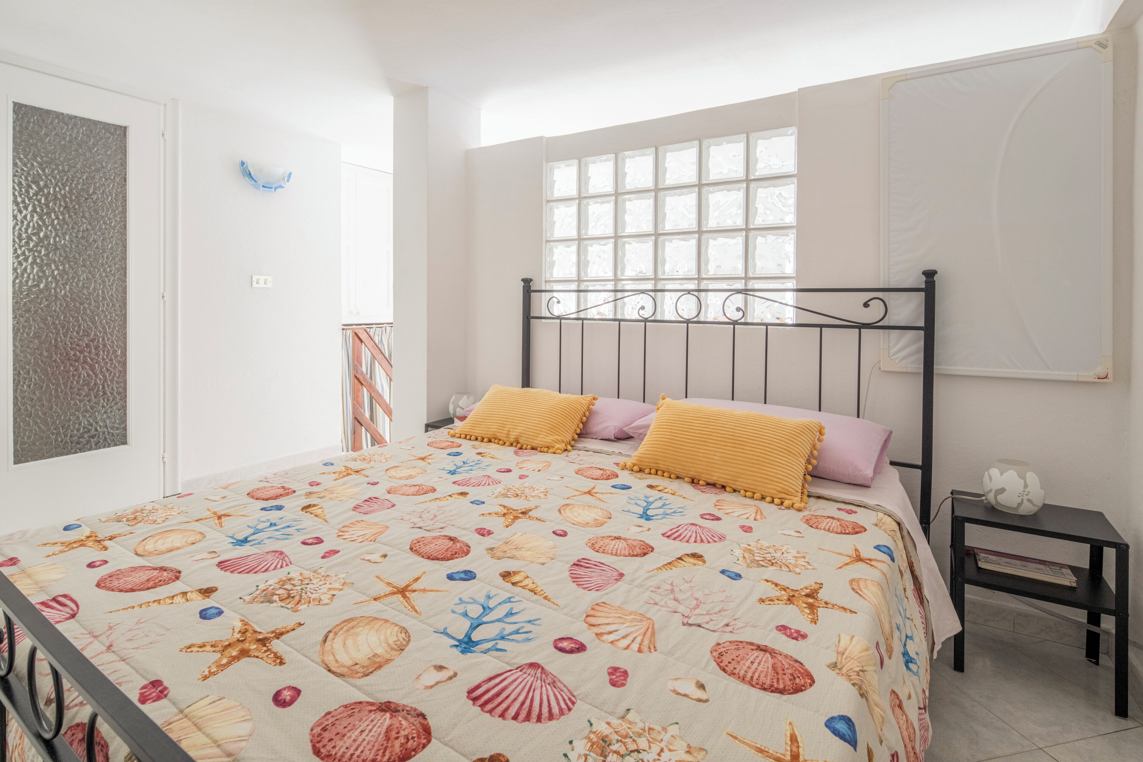 A bedroom with a bed featuring a seashell and starfish pattern, a window with blinds, a nightstand with a lamp, and a door with a textured panel.