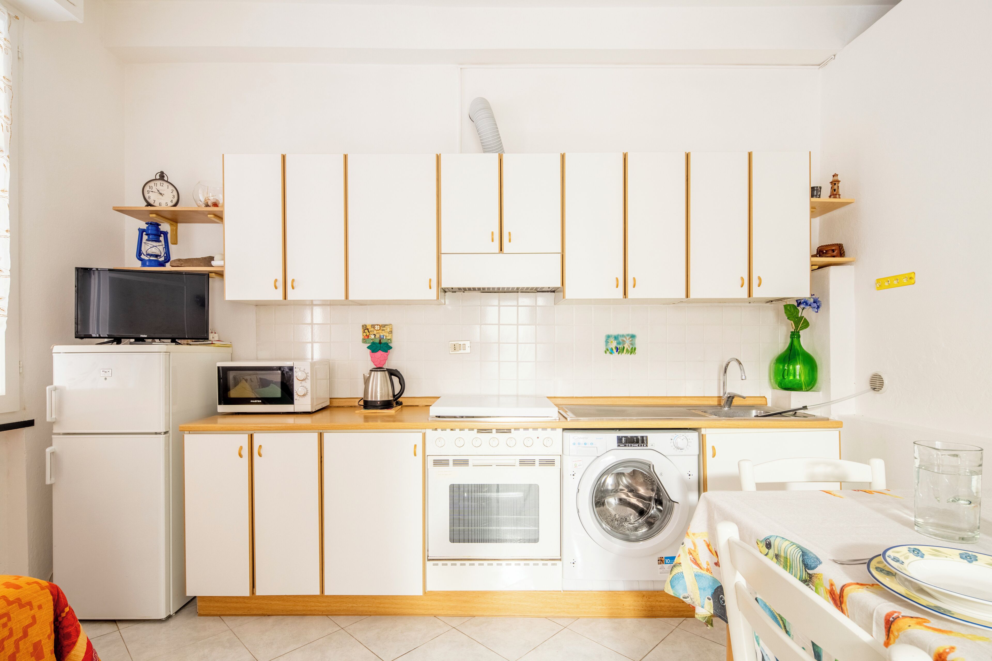 Una cocina compacta con muebles blancos, un microondas, un refrigerador, una lavadora y un lavamanos.