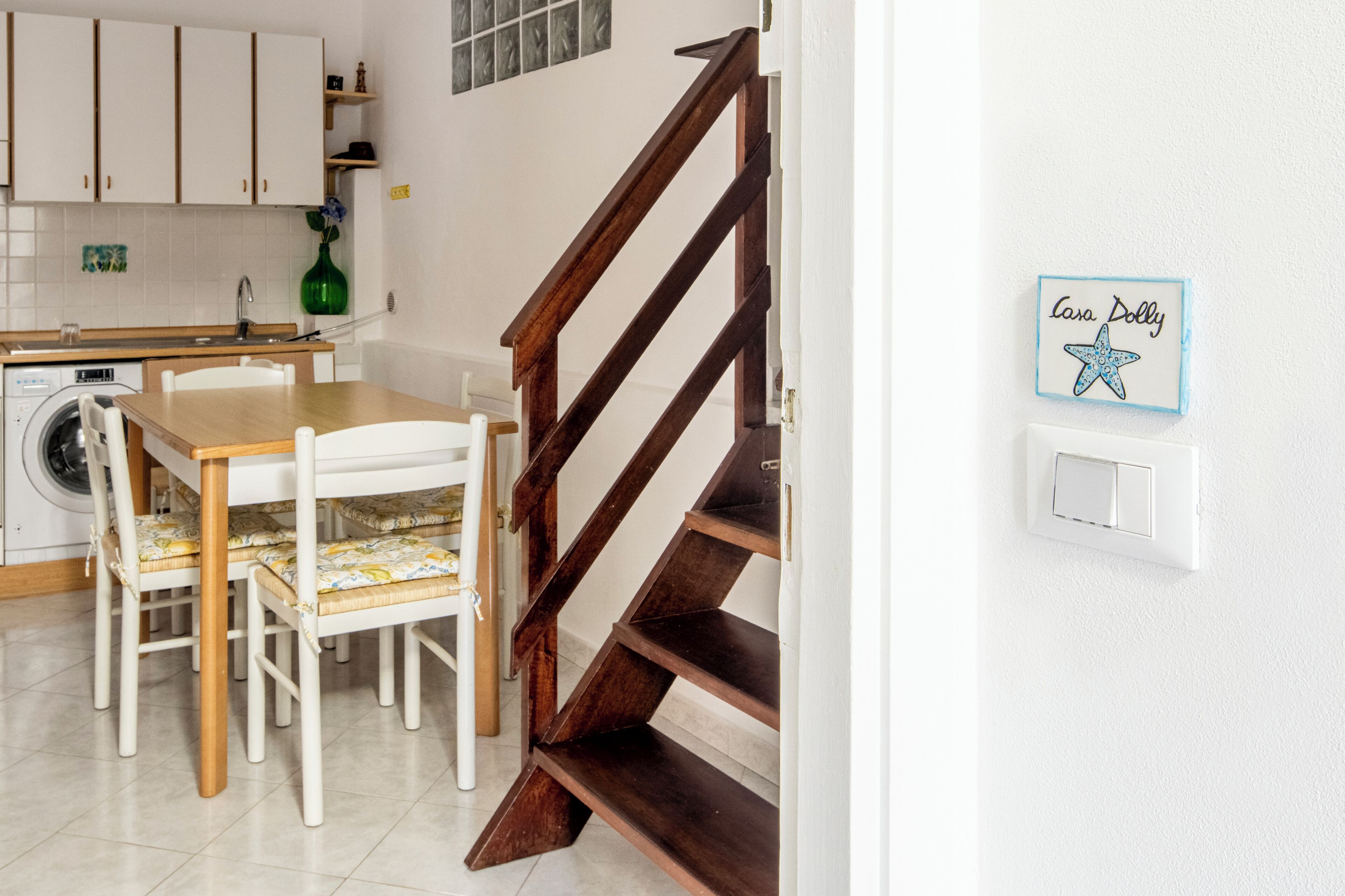 A kitchen with white cabinets, a wooden dining table with chairs, a washing machine, and a staircase.