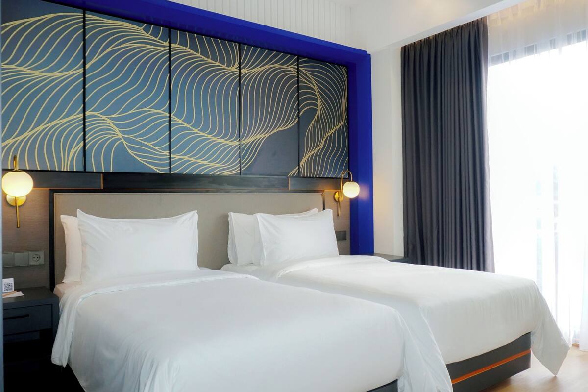 A hotel room with two beds, a modern headboard with a wave pattern, a bedside table with a lamp, and a large window with curtains.