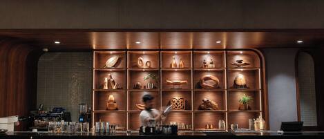 A bar area with a wooden shelf displaying various items, a bar counter with a coffee machine, and a person preparing a drink.
