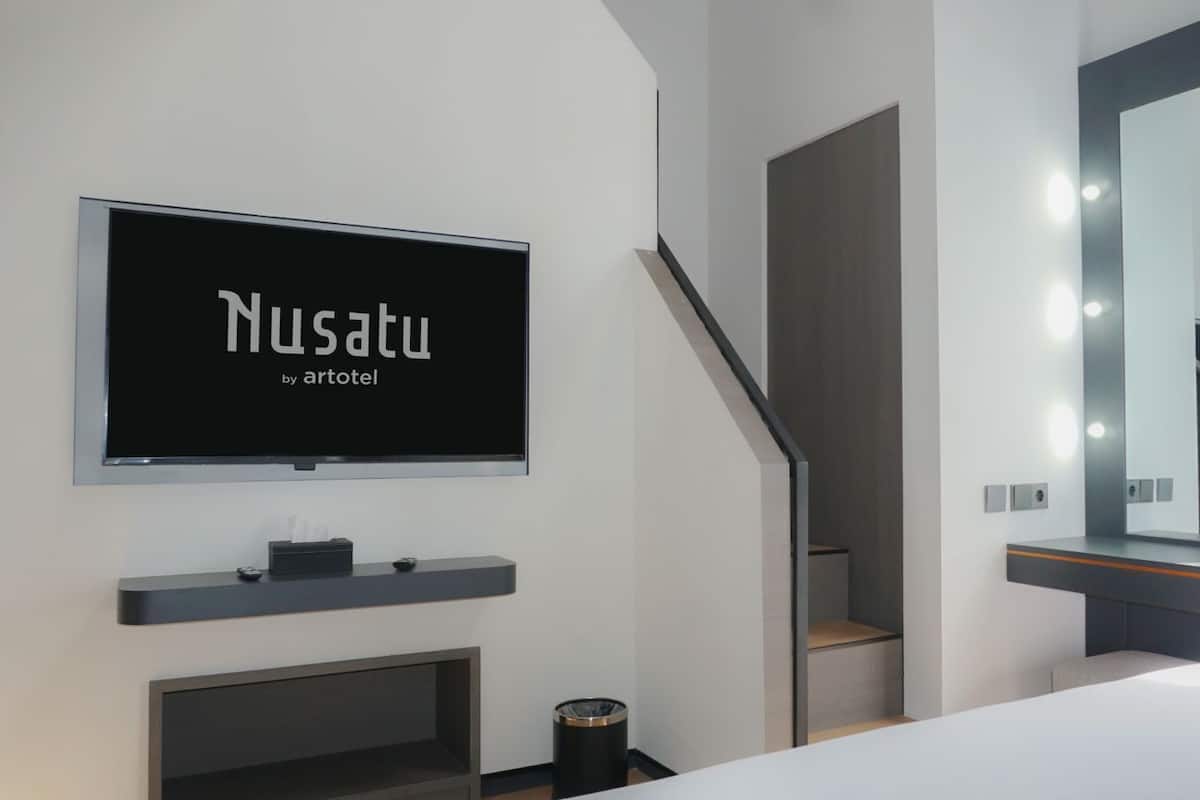 A modern hotel room with a large flat-screen TV mounted on the wall, a staircase, a vanity with a mirror, and a trash can.