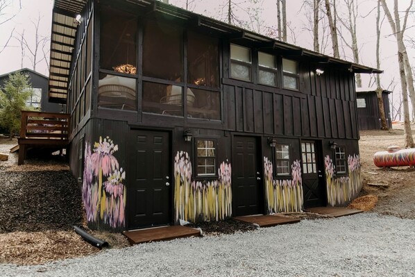A two-story wooden building with large windows and a door, surrounded by gravel and trees.