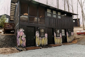 Ein zweistöckiges Holzhaus mit großen Fenstern und einer Tür, umgeben von Kies und Bäumen.
