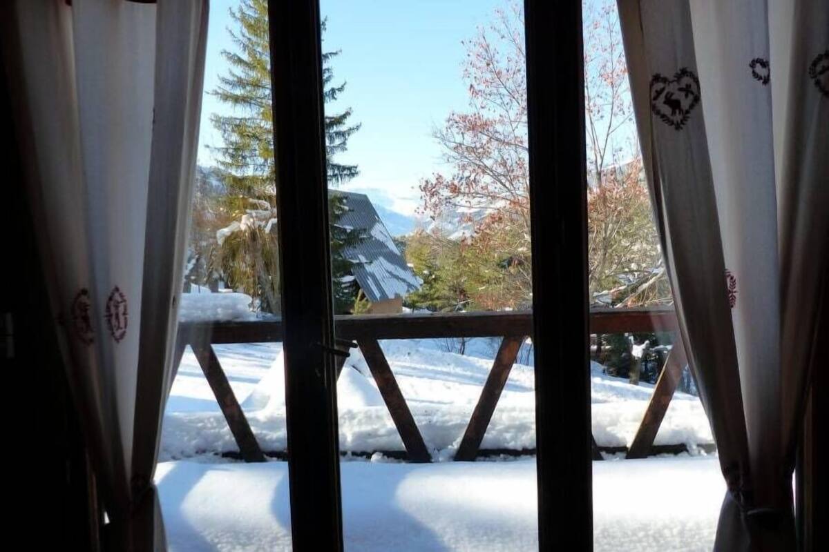 A view through a window with curtains, featuring a snowy landscape, a wooden deck, and a house with a sloped roof.