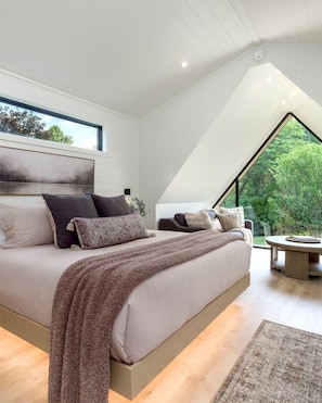 Een moderne slaapkamer met een groot bed, uitzicht op groen via een dakraam, en een ronde tafel met een stoel.