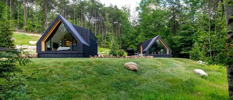 Two modern cabins in a forest setting with a grassy hill, surrounded by trees.