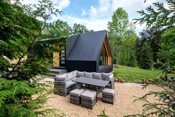 Une cabane moderne dotée d’un toit en pente, d’un espace extérieur aménagé pour s’asseoir et offrant une vue sur un paysage boisé.