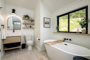 Een moderne badkamer met een vrijstaand bad, een toilet, een wastafel en een raam met uitzicht op groen.