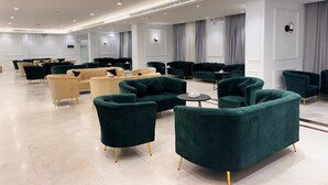 Un hall d’hôtel spacieux, doté de plusieurs espaces de détente, notamment des fauteuils vert foncé et des canapés beige.