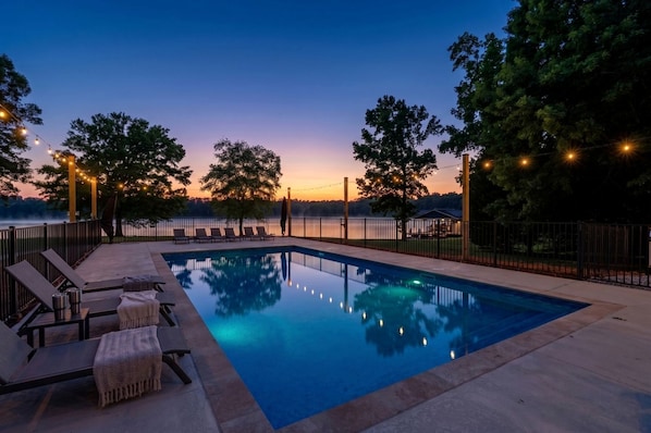 A rectangular pool with lounge chairs and a pergola, surrounded by a fence and trees, with a sunset in the background.