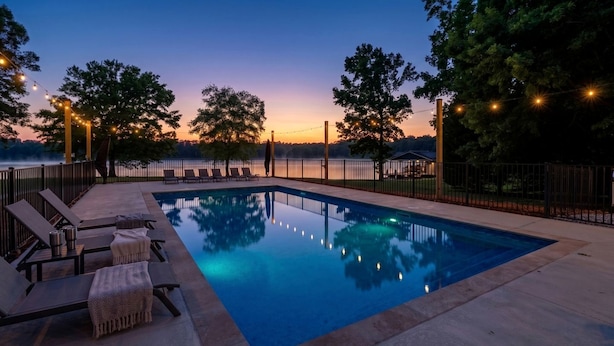 Una piscina rectangular con sillas de descanso y una pérgola, rodeada por una valla y árboles, con un atardecer al fondo.