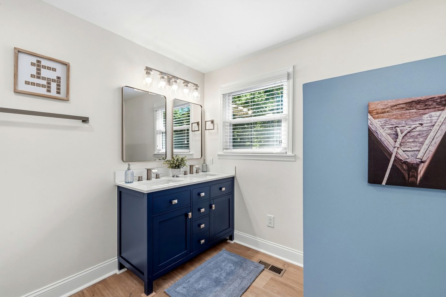 A bathroom with a double vanity, a large mirror with lights, a window with blinds, and a framed artwork on the wall.