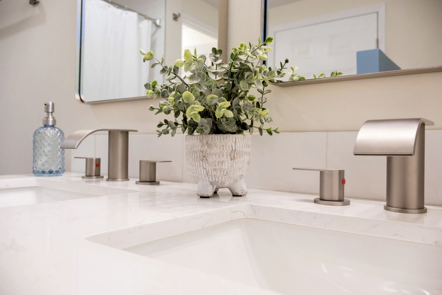 Une salle de bain avec un lavabo en marbre blanc, une plante dans un pot texturé, un distributeur de savon bleu et un robinet moderne.