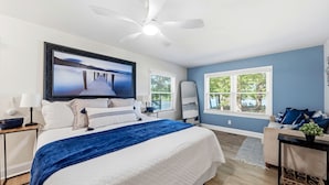Un dormitorio con cama, ventilador de techo, televisor, ventana con persianas y vistas al exterior.