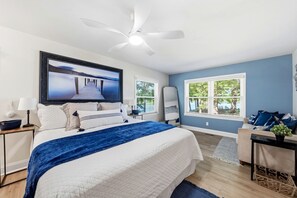 A bedroom with a bed, a ceiling fan, a TV, a window with blinds, and a view of greenery outside.