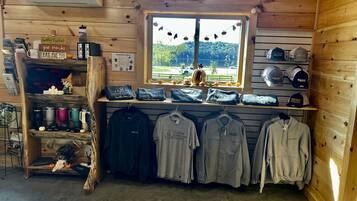 A rustic cabin interior with a wooden interior, a window with a view of trees, shelves with various items, and a display of T-shirts.