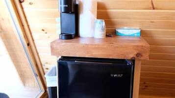 Un petit réfrigérateur noir orné du logo U-MAX, une machine à café, une tasse blanche et une boîte, le tout posé sur un comptoir en bois dans une cabane aux murs en bois et dotée d’une fenêtre.