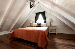 Une chambre avec un plafond en pente, un lit recouvert d’une couverture orange, deux tables de chevet avec des lampes et une fenêtre avec des rideaux noirs.