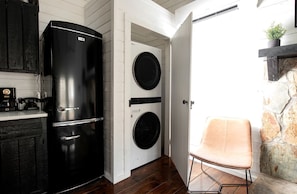Une cuisine compacte avec un réfrigérateur noir, deux machines à laver, un meuble en bois et une chaise.