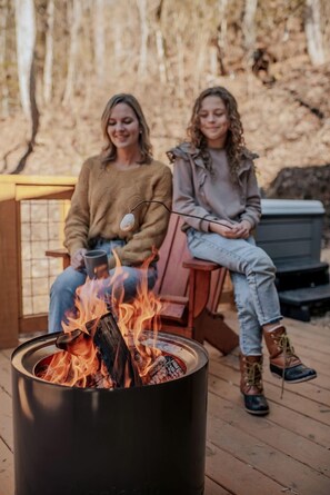 Two individuals seated by a fire pit on a wooden deck with a forest backdrop.