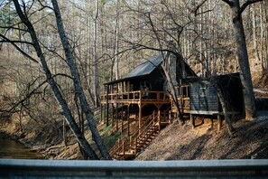 A cabin with a wooden deck and stairs leading up to it, surrounded by trees and a river.