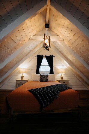 A bedroom with a sloped ceiling, a bed with a striped blanket, two bedside lamps, and a window with black curtains.