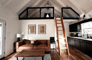 A loft-style room with a wooden floor, a brown leather sofa, a wooden coffee table, a ladder, a kitchen area with dark cabinets, and two framed pictures on the wall.