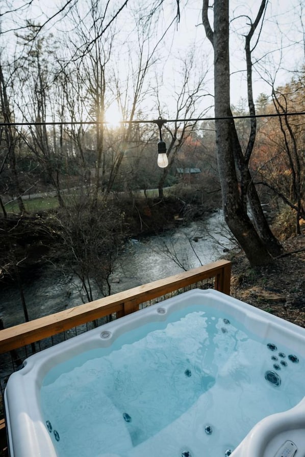 A hot tub with a wooden deck, surrounded by trees and a body of water.