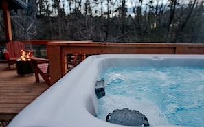 A hot tub on a wooden deck with a fire feature and chairs in the background.