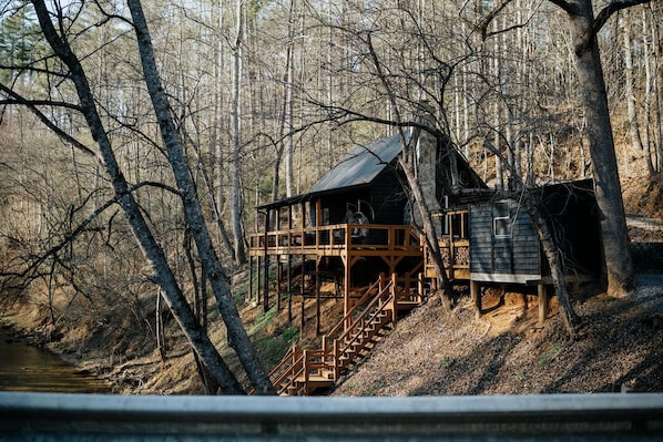 A cabin with a wooden deck and stairs leading up to it, surrounded by trees and a river.