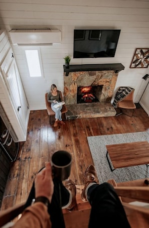 A room with a stone fireplace, a television mounted above it, and a person sitting on a chair reading a book.