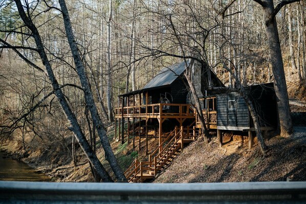 A cabin with a wooden deck and stairs leading up to it, surrounded by trees and a river.