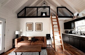 A loft-style room with a wooden floor, a brown leather sofa, a wooden coffee table, a ladder, a kitchen area with dark cabinets, and two framed pictures on the wall.
