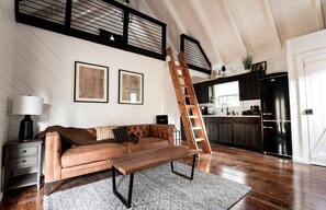 A loft-style living space with a loft bed, a wooden ladder, a sofa, a coffee table, a kitchen area, and framed artwork on the walls. - Hygge Hollow Cabin on Fightingtown Creek in Blue Ridge, GA (Blue Ridge)